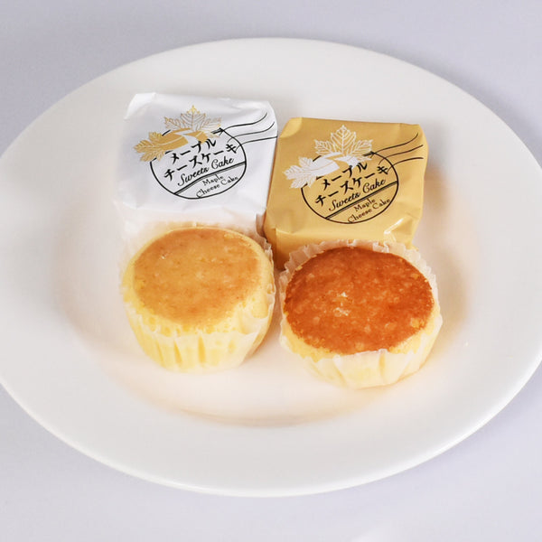 白い正方形の菓子箱の中身。白い丸皿に白の包装紙1個、ベージュの包装紙1個、丸いひとくちサイズのチーズケーキが2個載っている。
